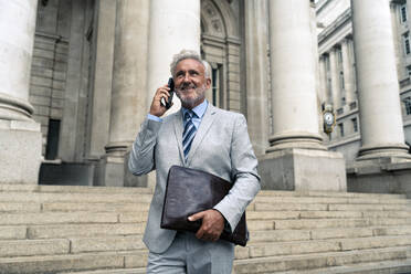 Smiling senior businessman talking on smart phone near building - OIPF03603