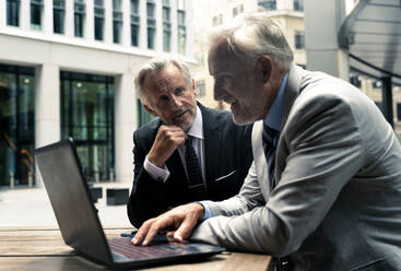 Lächelnder älterer Geschäftsmann, der mit einem Kollegen über einem Laptop am Tisch diskutiert - OIPF03641