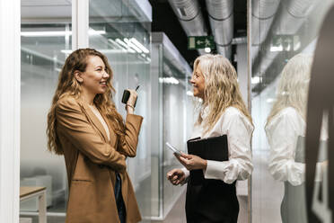 Smiling businesswomen holding smart phones and talking to each other in office corridor - PBTF00352