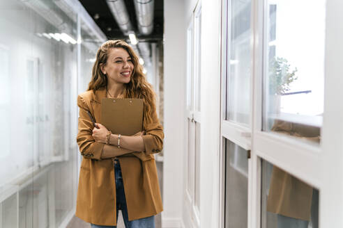 Lächelnde Geschäftsfrau mit Notizblock, die am Fenster im Büro steht - PBTF00358