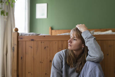 Contemplative woman with hand in hair sitting near bed at home - SVKF01686