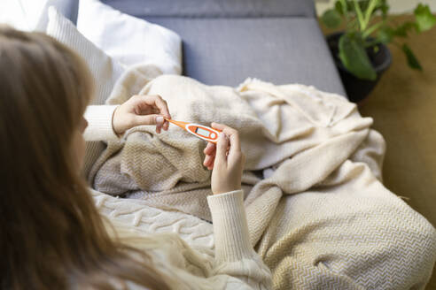 Frau überprüft digitales Thermometer auf dem Sofa sitzend zu Hause - SVKF01687