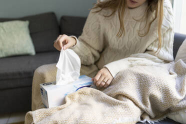 Woman pulling tissue paper from box sitting on sofa at home - SVKF01689