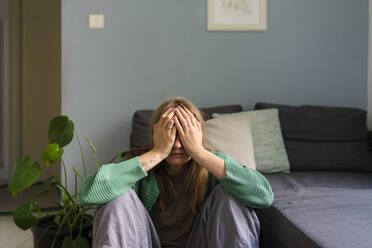 Stressed woman hiding face sitting near sofa at home - SVKF01698