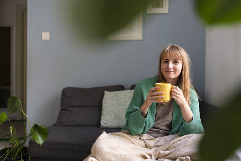 Lächelnde Frau, die eine Teetasse hält und zu Hause auf dem Sofa sitzt - SVKF01702