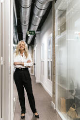 Confident blond businesswoman with arms crossed standing in office corridor - PBTF00387