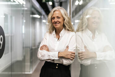 Blond businesswoman with arms crossed leaning on glass wall in office - PBTF00389