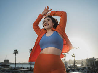 Happy woman in sports clothing standing with arms raised under sky - MFF09404