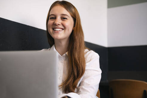 Lächelnde junge Frau mit Laptop im Büro sitzend - AMWF01937