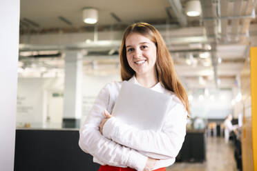 Smiling young businesswoman holding laptop at work place - AMWF01940