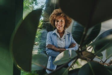 Smiling businesswoman standing with arms crossed behind plants near window in office - YTF01293