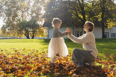 Lächelnde Mutter hält Hände mit Tochter in der Nähe von Herbstblättern vor dem Haus - NDEF01329