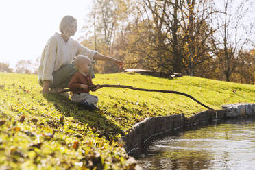 Mutter mit Sohn hält Stock in der Nähe des Sees an einem sonnigen Tag - NDEF01335