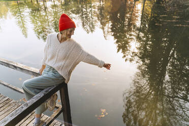 Smiling woman sitting with arm raised on railing near lake - NDEF01353