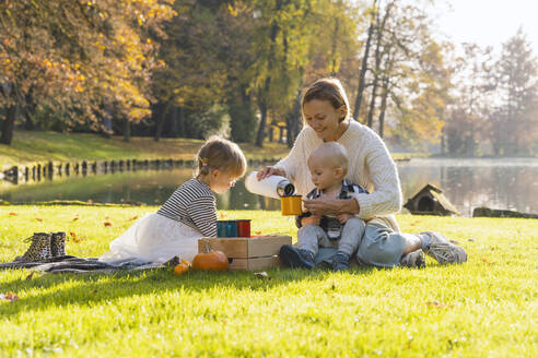 Glückliche Mutter gießt heiße Schokolade in eine Tasse mit Kindern, die am See sitzen - NDEF01359