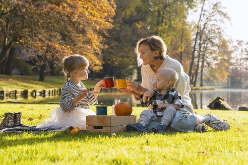 Lächelnde Mutter stößt mit ihrer Tochter in der Nähe eines Sees an einem sonnigen Tag auf eine Tasse an - NDEF01360