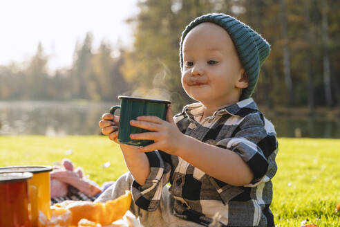 Lächelnder Junge mit Strickmütze, der beim Picknick eine Tasse heiße Schokolade hält - NDEF01363