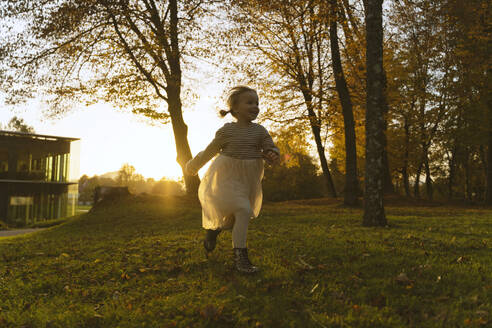 Glückliches Mädchen läuft in der Nähe von Baum im Park bei Sonnenuntergang - NDEF01368