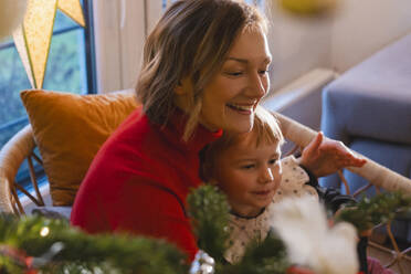 Smiling mother and daughter sitting on chair at home - NDEF01373