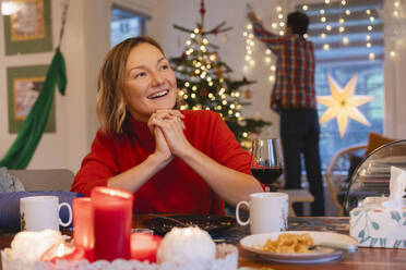 Glückliche Frau mit gefalteten Händen, die während der Weihnachtsfeier am Esstisch sitzt - NDEF01382