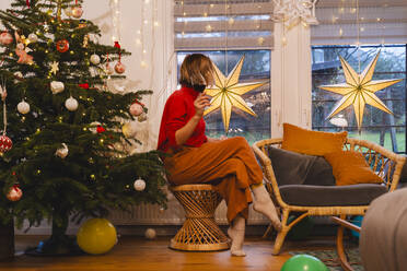 Frau mit rotem Weinglas, die während der Weihnachtszeit durch das Fenster ihres Hauses schaut - NDEF01386