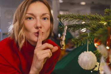 Blond woman gesturing finger on lips by Christmas tree at home - NDEF01390