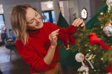 Lächelnde blonde Frau versucht rote Schleife am Weihnachtsbaum - NDEF01391