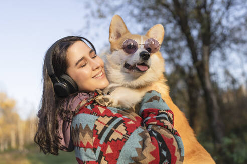 Happy woman carrying Pembroke welsh corgi wearing sunglasses in park - VBUF00484