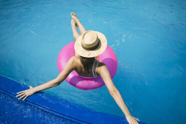 Woman wearing straw hat relaxing on inflatable swim ring in pool - YBF00281