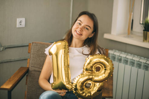 Smiling woman sitting on chair with golden number 18 balloons at home - ADF00235