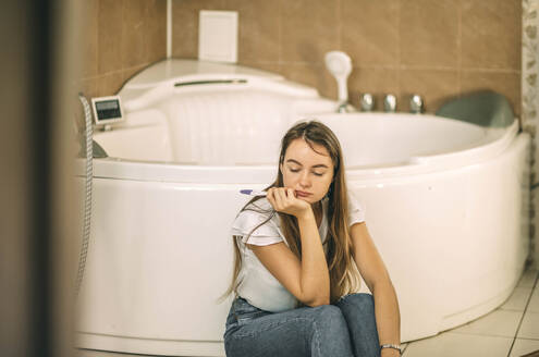 Traurige Frau hält Schwangerschaftstest-Kit und sitzt auf dem Boden im Badezimmer zu Hause - ADF00240