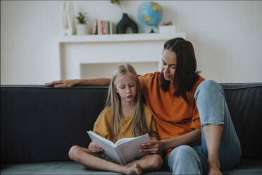 Mother and daughter reading book together on sofa at home - NLAF00181