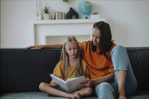 Mutter und Tochter lesen gemeinsam ein Buch auf dem Sofa zu Hause - NLAF00181