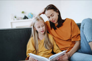 Girl reading book to mother on sofa in living room at home - NLAF00182