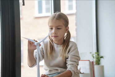 Blond girl playing with wind turbine model in window at home - NLAF00198