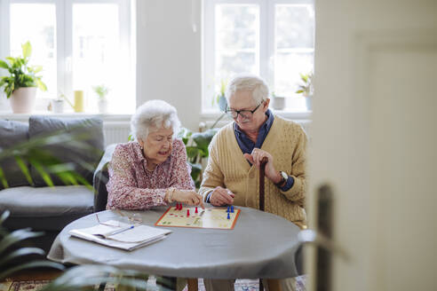 Senior woman playing ludo game with man at table in living room - HAPF03225