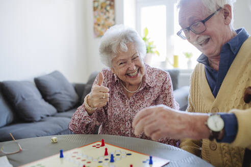 Cheerful woman gesturing thumbs up and playing ludo with man at table - HAPF03230