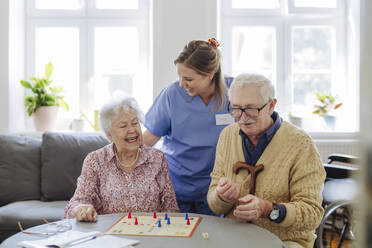 Glückliche Mitarbeiterin im Gesundheitswesen mit einem älteren Paar, das am Tisch im Wohnzimmer Ludo spielt - HAPF03233