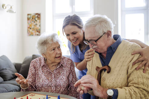 Lächelnde Pflegekraft mit älterem Paar, das zu Hause ein Ludo-Spiel spielt - HAPF03235