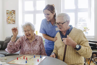 Glückliches älteres Ehepaar spielt Ludo mit einer Pflegekraft, die zu Hause steht - HAPF03236