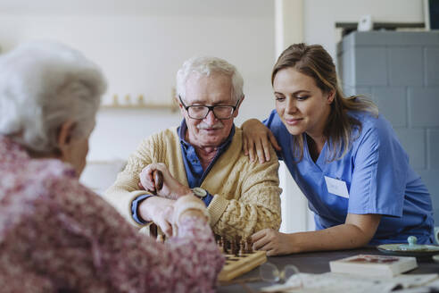 Smiling healthcare worker playing chess with senior couple at home - HAPF03238