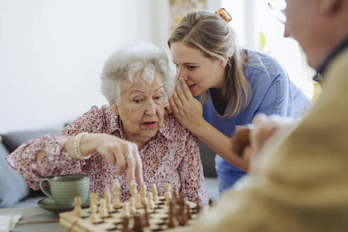 Pflegekraft flüstert einer älteren Frau ins Ohr und spielt zu Hause Schach - HAPF03242