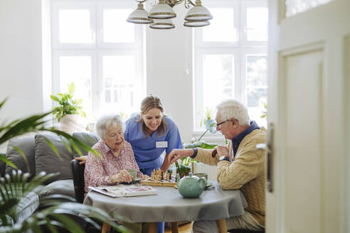 Smiling healthcare worker with senior couple playing chess at table - HAPF03243