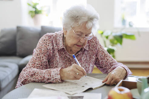 Senior woman with pen doing crossword puzzle in book at table - HAPF03249