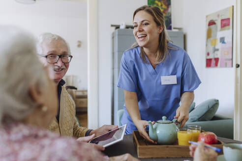 Happy healthcare worker holding teapot and talking to senior couple at table - HAPF03256