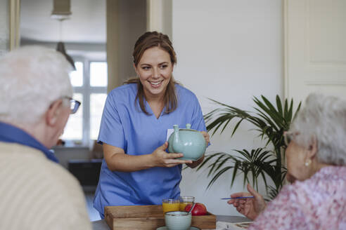 Happy healthcare worker holding teapot and talking to senior couple at home - HAPF03259