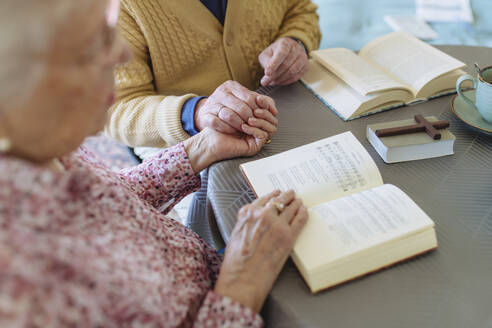 Senior couple holding hands and reading book at table - HAPF03263