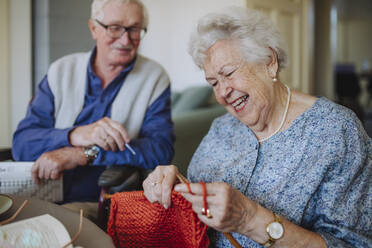 Smiling senior woman knitting with man sitting in background - HAPF03267