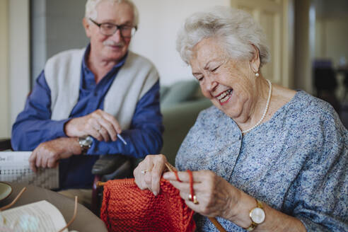 Lächelnde ältere Frau beim Stricken mit einem Mann im Hintergrund - HAPF03267