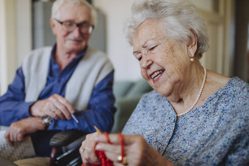 Happy senior woman knitting with man sitting in background - HAPF03268